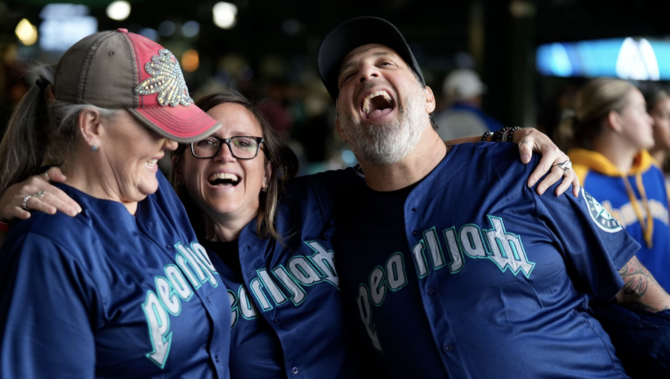 Three people in Mariners jerseys that say "Pearl Jam" laugh at T-Mobile Park during a Mariners game.